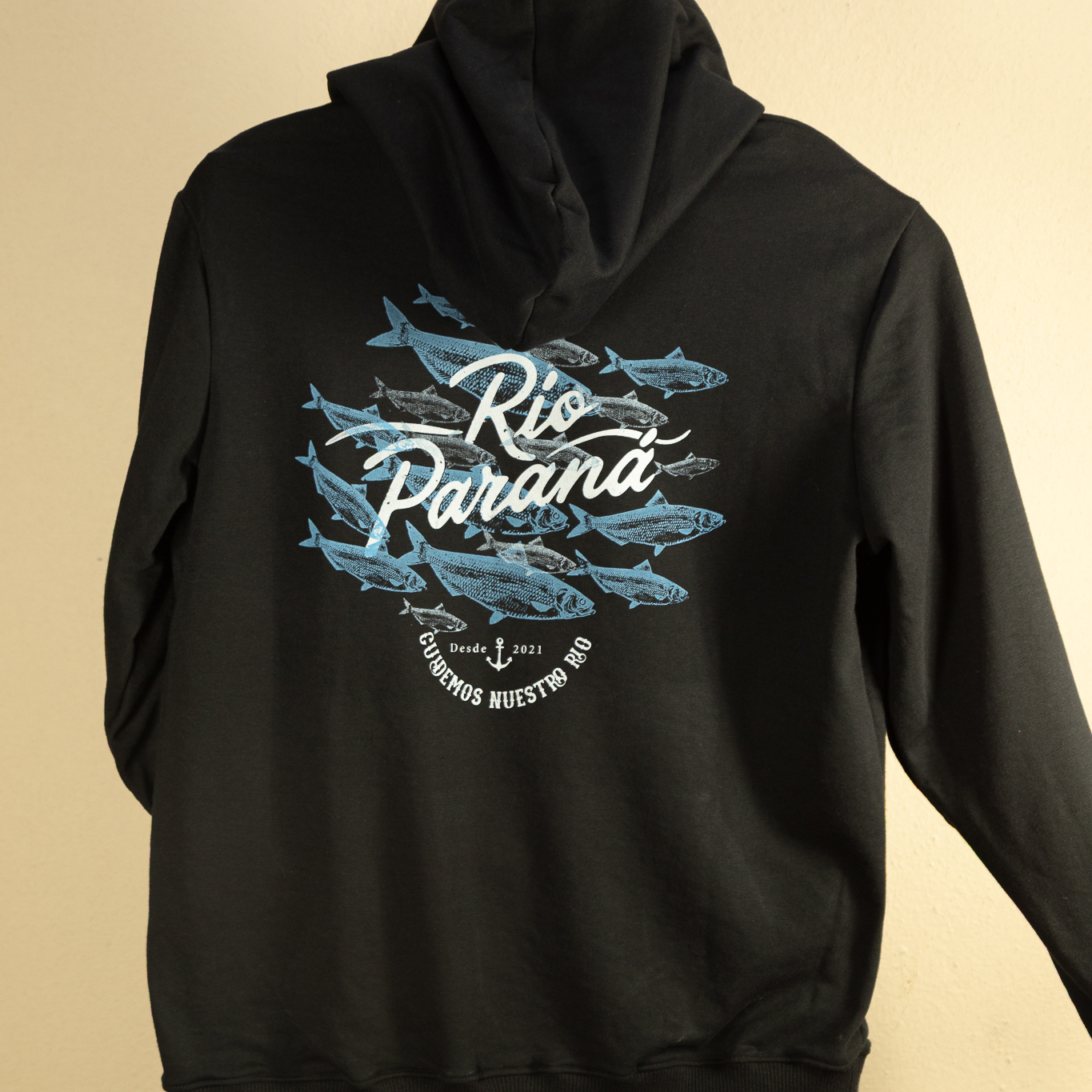 Buzo negro con capucha y canguro en rústico invisible peinado. Estampa dorso serigrafía artesanal: cardumen de sábalos en azul, firma Río Paraná en script blanco, ancla y texto Cuidemos Nuestro Río. Hecho a mano en Corrientes. Marca Río Paraná desde 2021.